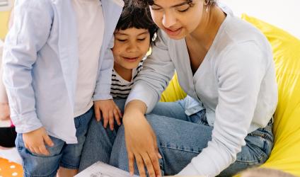 A teacher sitting in a low chair with 3 young kids gathered around and all are looking at an open book