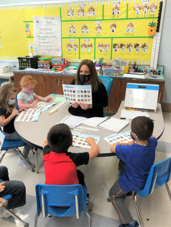 [image of a teacher sitting in a classroom holding up a book towards young students seated at a semi-circle table around her]